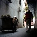 Elderly man carrying bags in a narrow urban street with a conical hat in a cart.